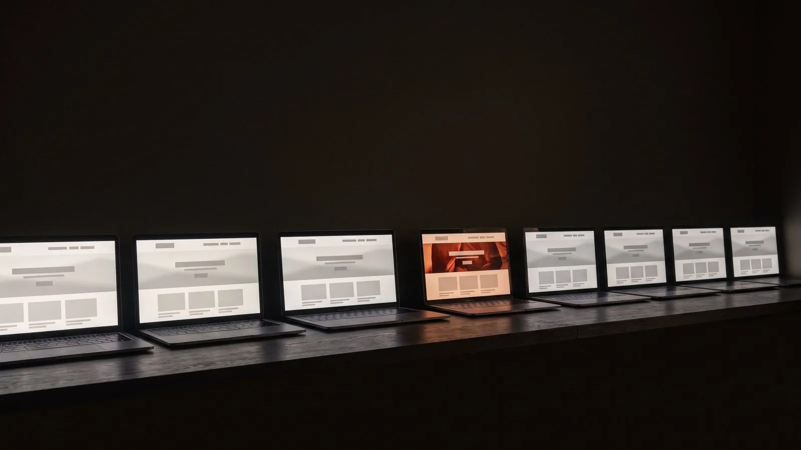 Nine laptops in a row on a dark shelf, eight showing the same wireframe homepage, one showing a page built around a photograph